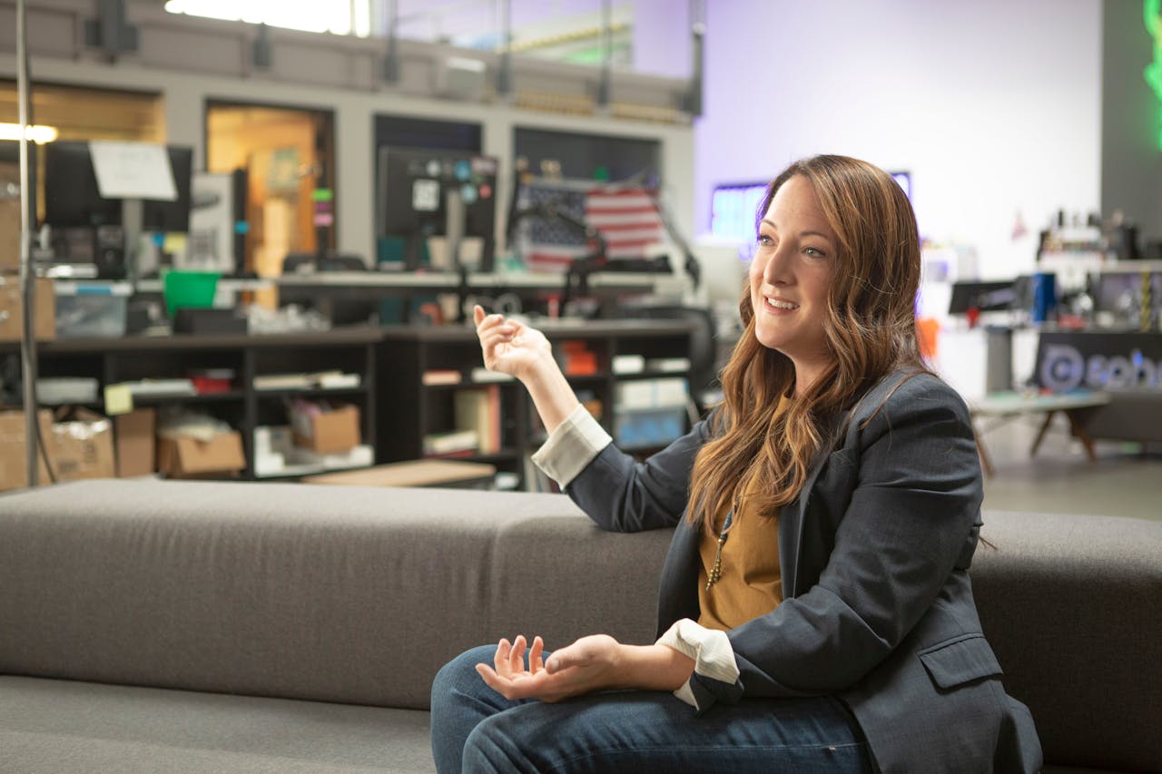 Businesswoman speaking in a modern office setting, wearing a blazer and sitting on a couch.