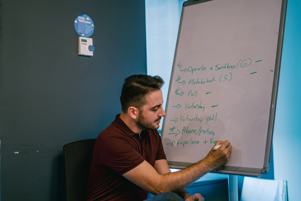 Bearded man in a polo shirt writing on a whiteboard with markers in an office setting.